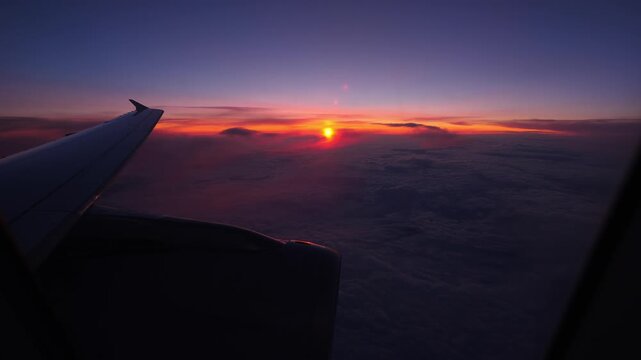 Dramatic sunset view from airliner window, red sun hang low and almost hide, colored stripe at horizon line, clear sky above and dark clouds seen down. Plane wing and engine on foreground