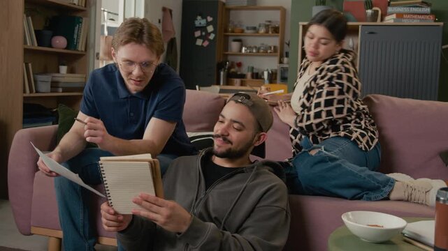Diverse young zoomers discussing notes in notebook while studying for exams together in living room. Higher education marketing, student lifestyle promos, collaborative learning concepts