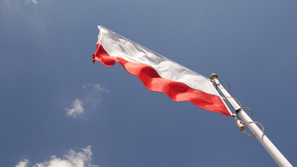 Biało-czerwona flaga państwowa powiewa dynamicznie na wietrze, zamocowana na ukośnym metalowym maszcie. W tle widoczne jest głębokie, niebieskie niebo z kilkoma małymi, białymi chmurami. © tomo81