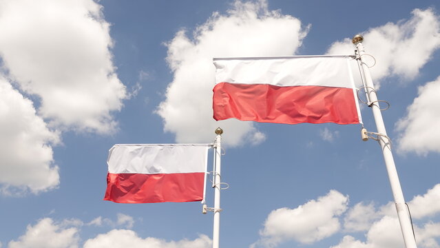 Dwie biało-czerwone flagi Polski powiewające na masztach na tle błękitnego nieba z białymi, puszystymi chmurami. Narodowe symbole uchwycone w pełnym słońcu symbolizują patriotyzm i wolność