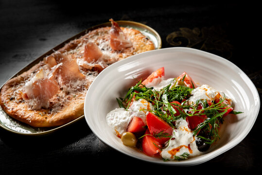 Gourmet flatbread with prosciutto and a fresh tomato salad served with stracciatella cheese on a dark background.