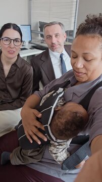 Portrait of African American woman holding sleeping baby in carrier, talking to camera with Caucasian colleagues in office. Clip representing working parents and diversity, vertical
