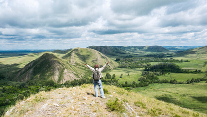 Fototapeta premium Alone backpacker woman stands on a rocky hillside overlooking a wide green valley with rolling mountains, scattered trees, and a cloudy sky in the background