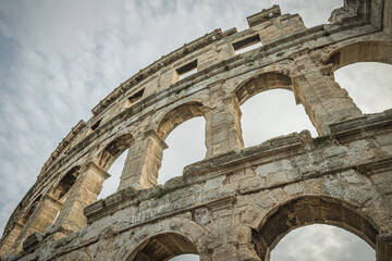 Obraz premium The ancient Roman amphitheater in Pula, Croatia, showcasing its impressive stone arches and architectural details against a cloudy sky. Pula, Croatia.