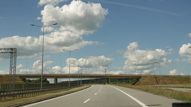Szeroka, pusta autostrada prowadzi w kierunku betonowego wiaduktu przecinającego trasę. Nad drogą rozpościera się błękitne niebo wypełnione dużymi, malowniczymi białymi chmurami.