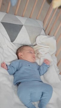 Newborn hispanic baby boy sleeping on white blanket in wooden crib wearing blue knit romper, curled hand near cheek in building; serene rest.