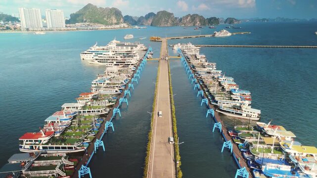 Halong bay marina lined with tour boats and cruise ships along a long pier, gateway to limestone karsts and clear blue waters with coastal buildings in the background