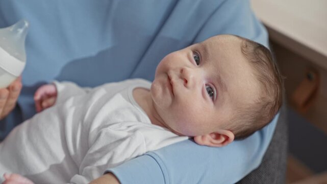 Newborn boy drinking from bottle, mouth latched and caregiver hand holding bottle while resting on caregiver lap in home; calm bonding.