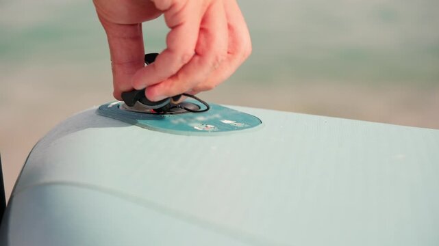 Close-up view of a man's hand pressing the valve to deflate an inflatable stand up paddleboard after a session, with the ocean waves gently rolling in the background on a sunny day