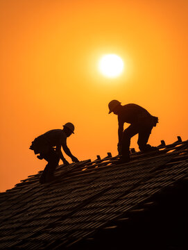 Silhouette of two construction workers repairing roof at sunset, male roofers installing shingles on house top against bright orange sun, industrial building service concept photography