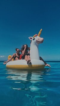 Kids playing with inflatable unicorn in turquoise ocean as float tips and splashes, two women guiding two boys, energetic summer fun, close water perspective, bright blue sky, vertical video
