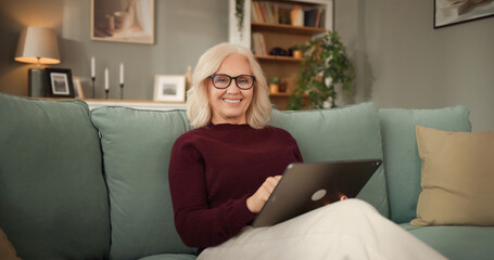 A woman sits on a couch in a living room, smiling as she uses a tablet. The room has bookshelves and plants, creating a cozy atmosphere in the evening. © Stockphotodirectors
