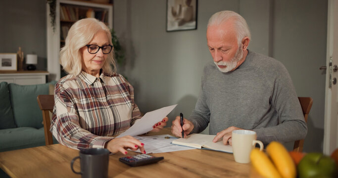 An older couple sits at a table in their home, reviewing life insurance papers. One person writes notes while the other holds a document and looks on.