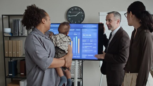 African American woman holding baby while discussing financial charts on screen with Caucasian colleagues in office. Ideal for promoting workplace diversity and supportive corporate culture