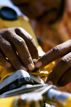 Surveying instrument close up of hand adjusting fine screw on yellow survey equipment under bright daylight, showing skin texture and metal detail