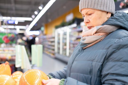 Elderly female customer wearing scarf choosing groceries while standing with trolley in contemporary retail store interior.