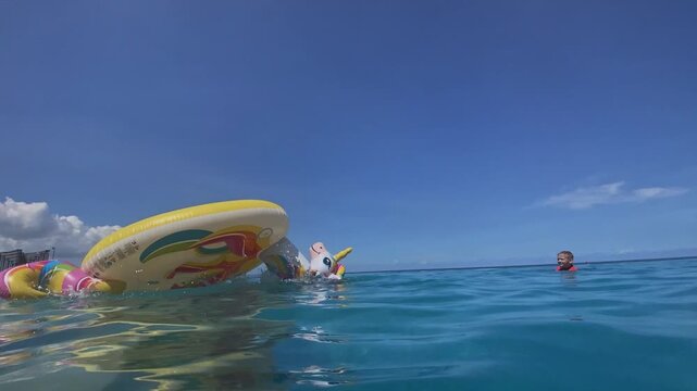 Caucasian boy on unicorn float drifting across calm turquoise ocean under clear blue sky, reflective water surface, distant clouds, slow ripples, relaxed summer leisure, Bali island