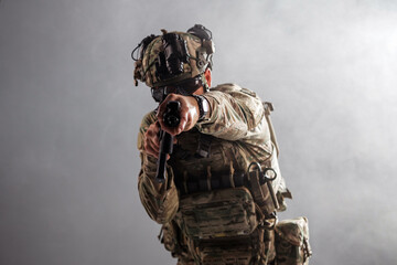 Obraz premium Armed soldier in tactical gear aiming firearm towards the camera, wearing camouflage uniform and protective equipment, with a smoky background creating a dramatic effect