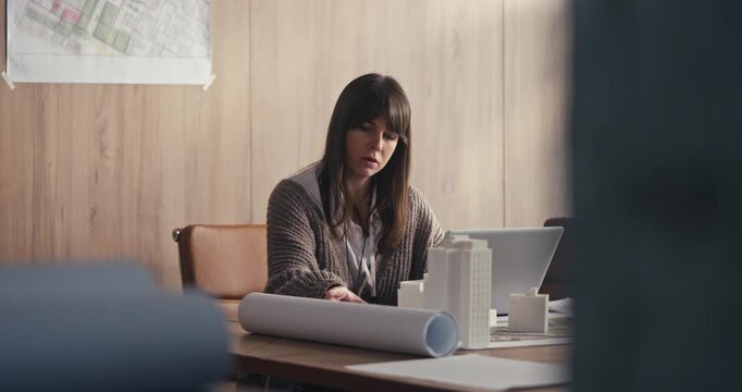 A woman draws and reviews architectural plans while sitting at her desk in an office setting during the late afternoon