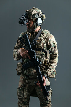 Male soldier in camouflage tactical gear holds a rifle while wearing a helmet with night vision goggles against a gray background