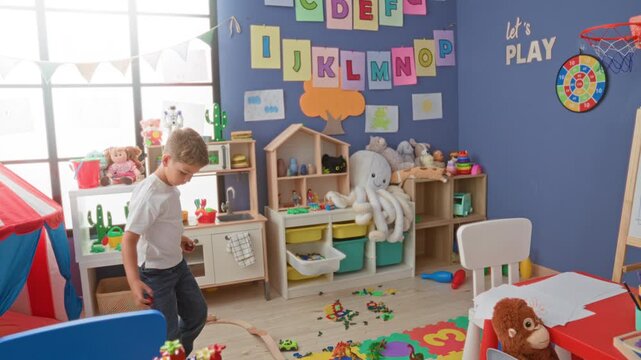 Boy throws a small red ball with his right hand inside a colorful children's playroom building scattered with blocks, tent and stuffed toys; playful joy.