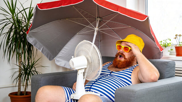 Man relaxes under an umbrella indoors while sitting on a couch with a fan nearby during summer heat