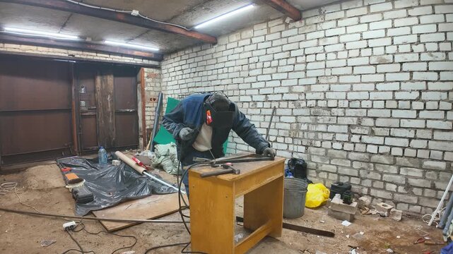 Industrial worker wearing a protective mask welding metal parts in a messy garage, with bright sparks flying from the electrode during the fabrication process in the workshop
