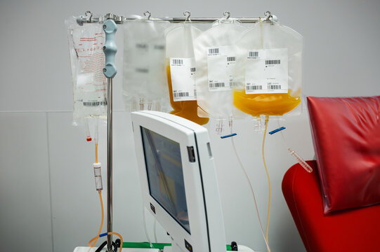 Plasma bags hanging on an IV stand beside an apheresis machine and red donation chair at a blood bank illustrating plasma collection and blood component donation.
