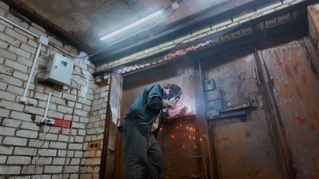 Low-angle view of a skilled industrial worker in a protective mask performing welding on a heavy-duty steel door inside a gritty workshop, with bright sparks flying from the intense heat