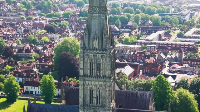 Aerial drone shot of Salisbury Cathedral in Wiltshire, England, showing the medieval Gothic cathedral spire, historic stone architecture, city rooftops, trees and surrounding urban landscape on a brig