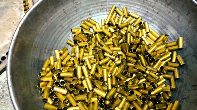 Golden brass bullet casings being sorted and agitated in a large metal bowl on an industrial production line at an ammunition factory, showcasing the manufacturing process of military ordnance