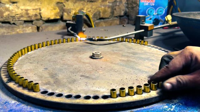 Close up view of a factory worker's hand placing bullet shells into a machine for heat treatment, showcasing the manual labor involved in ammunition and firearm cartridge manufacturing