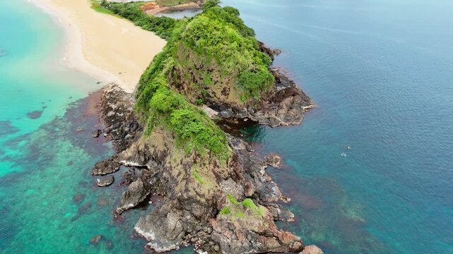 Aerial drone view of a rocky vegetated headland jutting into turquoise and dark blue water with a sandy beach adjacent and two outrigger boats nearby, Nacpan Beach, El Nido, Palawan, Philippines.