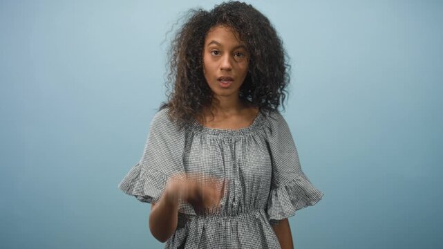 Teen woman with curly hair points finger forward and purses lips while standing against a blue studio backdrop, holding an index gesture and a contemplative pause; confidence.