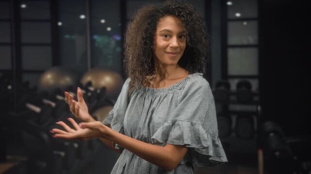 Woman gesturing with hands toward dumbbells in gym, smiling and showing bare arms and curly hair while wearing a gingham dress; confidence wellness positivity.