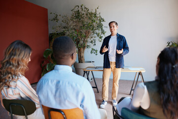 Fototapeta premium Young male presenter leading a business workshop, actively speaking and gesturing to a diverse group of attentive attendees during an educational seminar or corporate training session