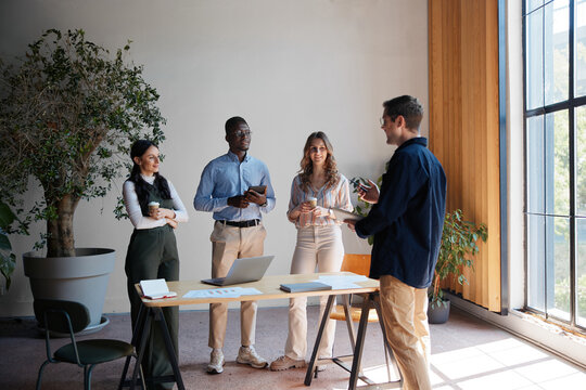 Diverse business professionals conducting a stand-up meeting, reviewing documents and sharing ideas, fostering teamwork and collaboration in a bright, contemporary workspace with natural light