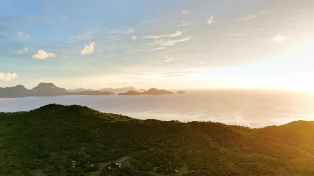 High-altitude aerial view of a coastal bay with forested hills, a winding road, village structures and limestone islands at golden sunset, Barotuan, El Nido, Palawan, Philippines.