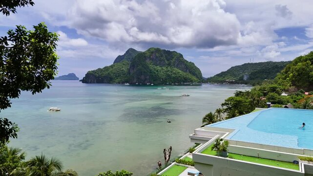 Aerial drone view of an infinity pool overlooking El Nido bay with a large jungle-covered limestone karst peak, moored boats and storm clouds, El Nido, Palawan, Philippines.