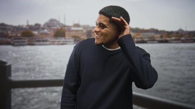 Hispanic man with beard cupping hand to ear while smiling on street by a waterfront railing; playful curiosity.