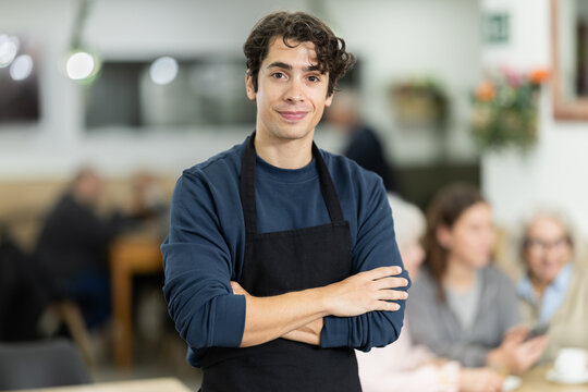 Young positive polite waiter guy posing in small coffee shop