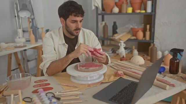 Man with beard shapes clay vessel with hands and small tool at pottery wheel in studio, surrounded by tools and pots; concentration craft.