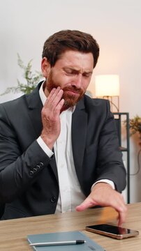 Middle-aged businessman at home office drinks cold water feels sharp tooth pain and searches dentist on smartphone. Freelancer guy at table presses cheek sighs hoping to book urgent clinic visit soon.