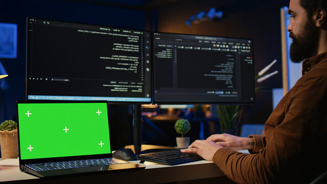 Programmer coding on computer next to isolated screen laptop in home office, developing software application. Teleworking IT specialist doing programming on PC near chroma key notebook.