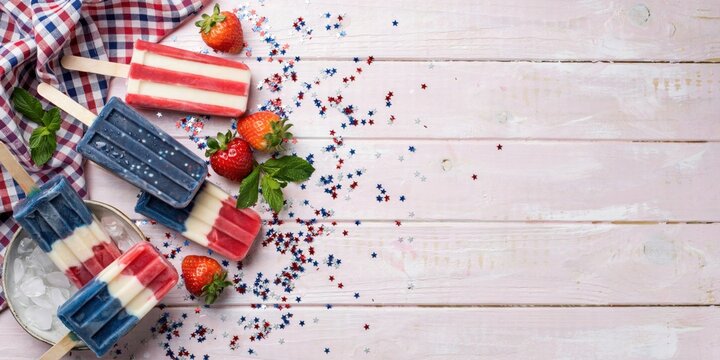 Popsicles with red white and blue layers sit on a rustic wooden table alongside fresh strawberries and mint leaves.