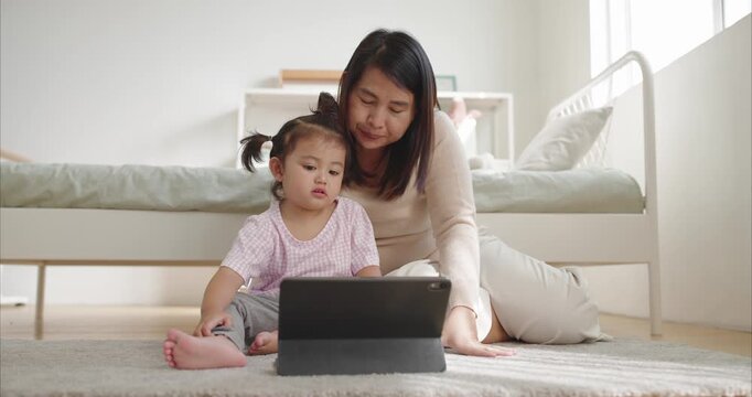 Asian Mother and little Girl Watching Tablet on Floor together morning light in bedroom, Concept digital parenting