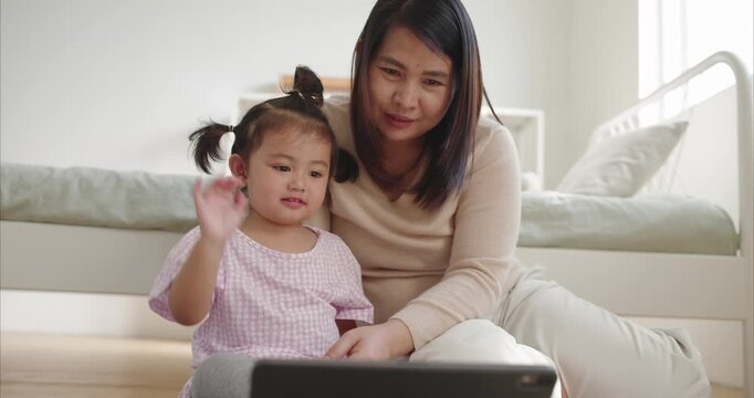 Asian Mother and Two Year Old Toddler Girl Watching Tablet on Floor,  Happy Mom and Child Using Digital Device Together in Cozy White Bedroom, Online Education and Family Quality Time