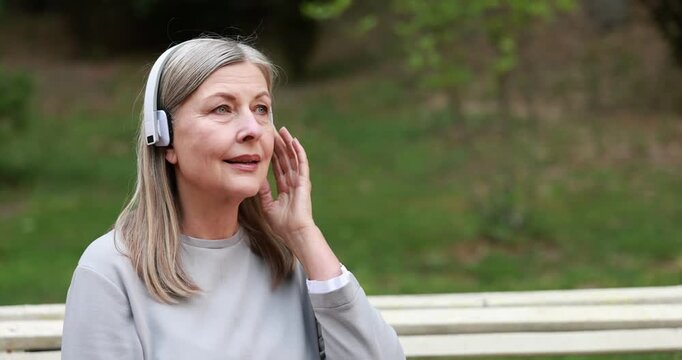 Beautiful senior woman with headphones sitting on bench in spring park, space for text. Camera moving forward