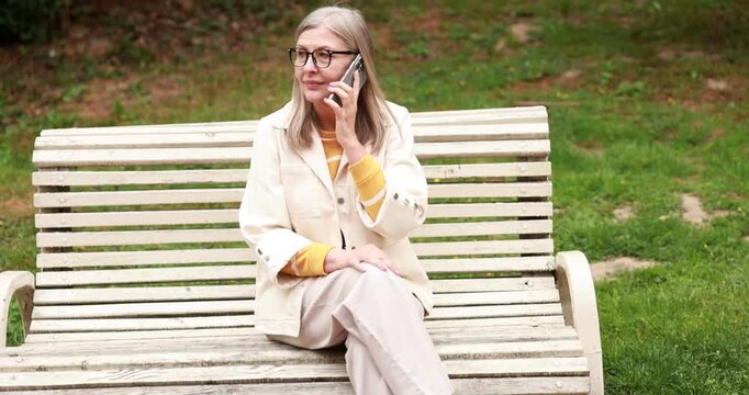 Beautiful senior woman talking on phone in spring park. Camera moving forward