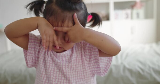 Sleepy Asian Toddler Girl Rubbing Eyes in Minimalist Room; Two Year Old Child Tired After Playing, Looking Drowsy in Cute Medium Shot Portrait, Childhood Fatigue and Bedtime Routine.